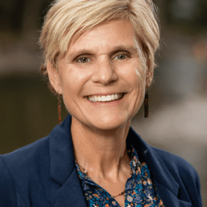 A photo of Laurel Angell, who is smiling and has shorter blonde hair, and is wearing a dark blue blazer and a blue and multi-colored blouse.