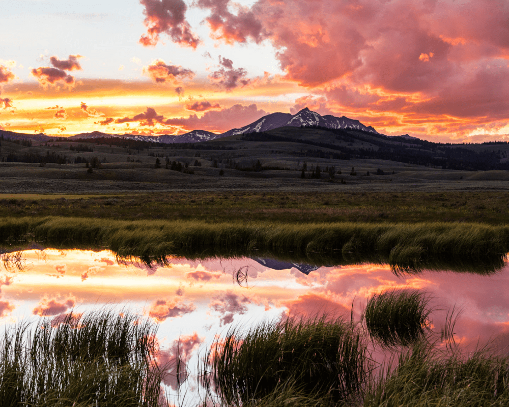 Behind a mountain in the distance is a vibrant pink, yellow and orange sunset on a cloudy evening, reflecting off a small body of water in the foreground.