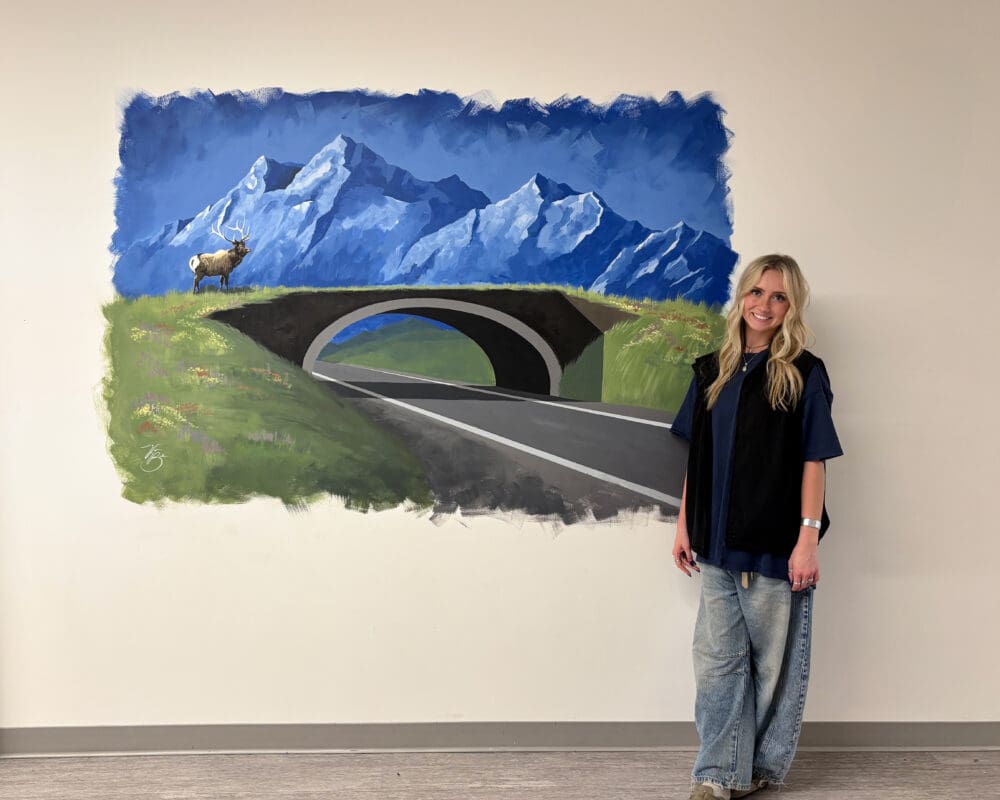 A student stands in front of her mural of a wildlife crossing in a Bozeman, Montana high school.